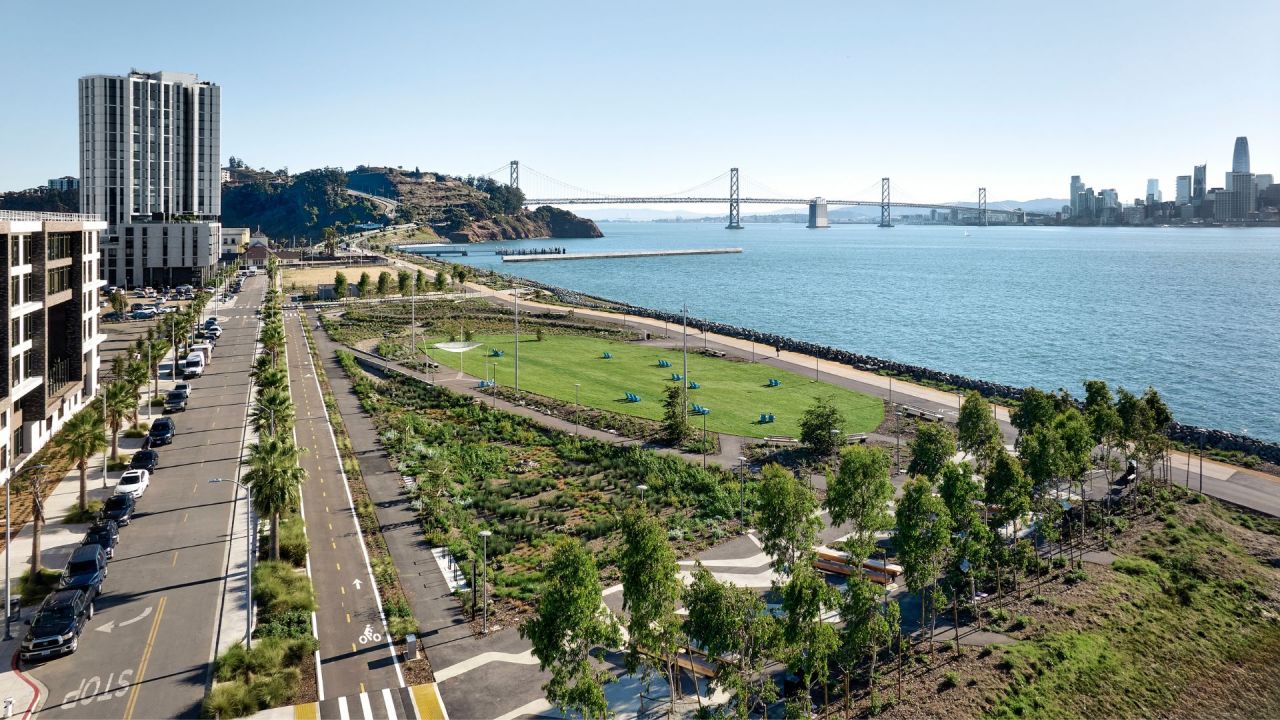 A tranquil path leads to a distant city skyline under a cloudy sky, with a bridge on the left.