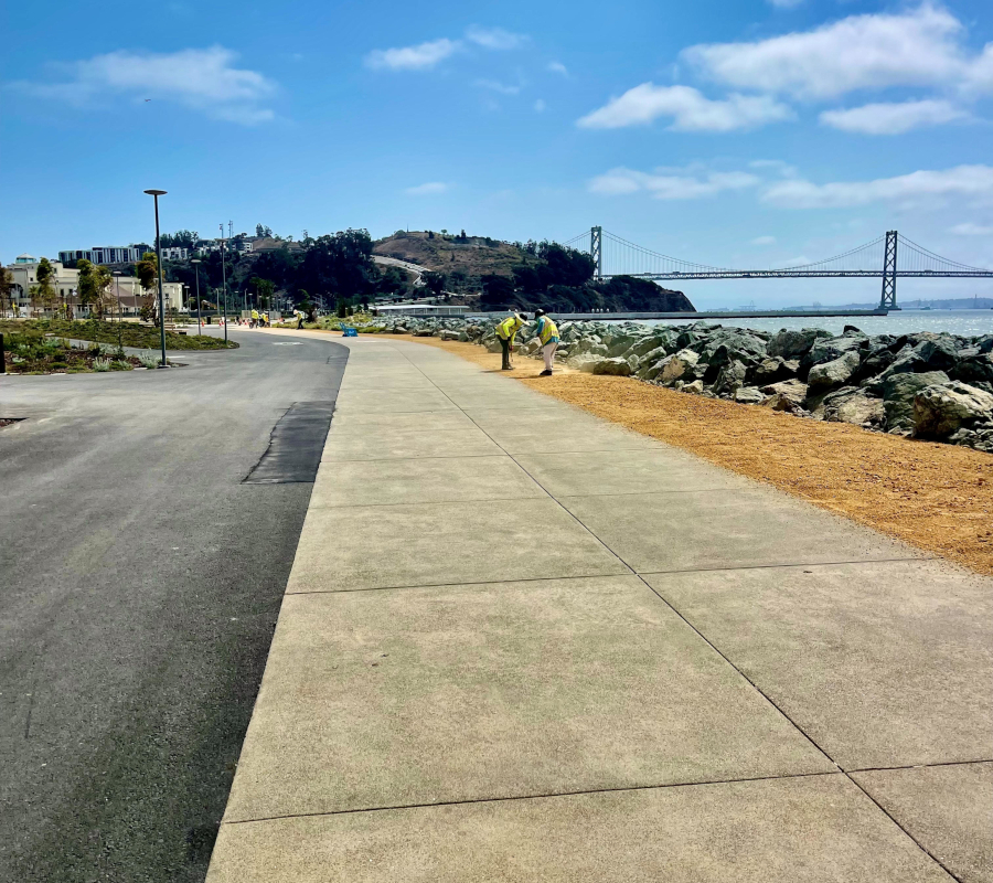 A scenic view of a coastal walkway beside a road, bordered by rocks and the sea. In the distance, two people walk under a partly cloudy sky near a large bridge.