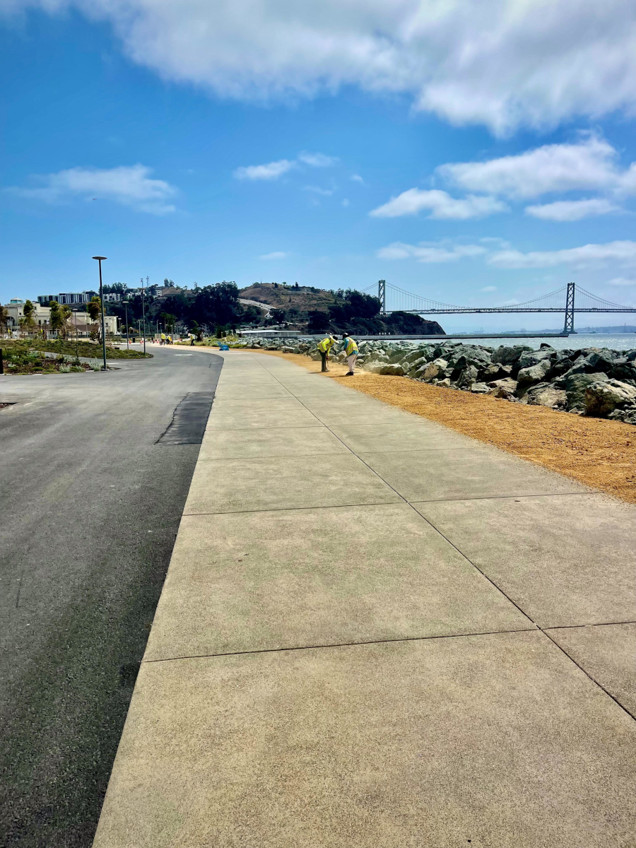 A scenic view of a coastal walkway beside a road, bordered by rocks and the sea. In the distance, two people walk under a partly cloudy sky near a large bridge.