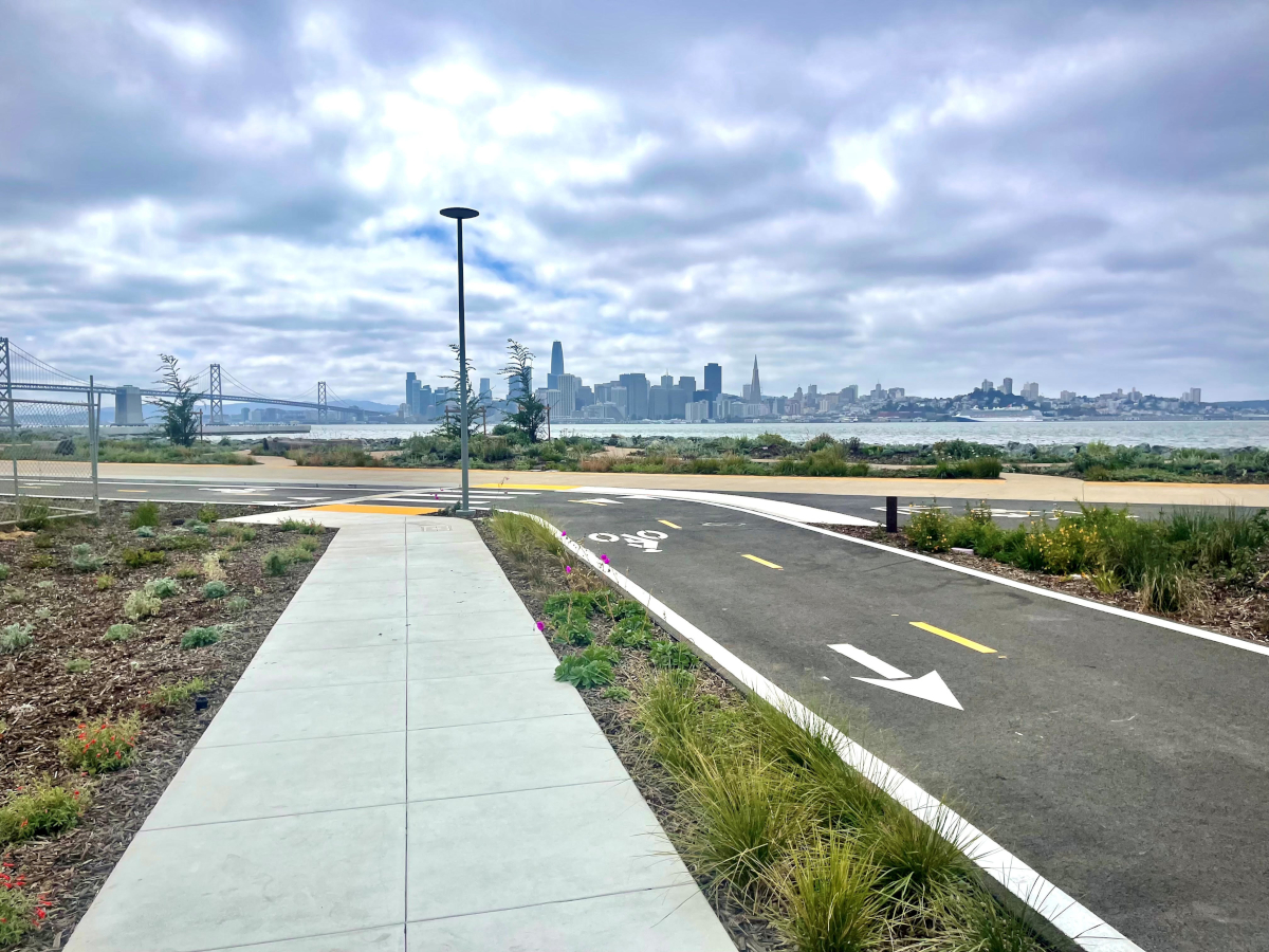 A tranquil path leads to a distant city skyline under a cloudy sky, with a bridge on the left.